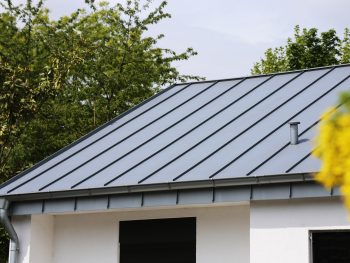 A residential house features a durable, light gray aluminum roofing with standing seams under a bright sky.