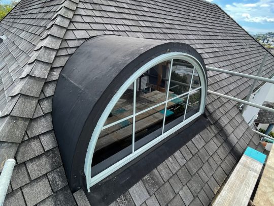 Architectural roofing: Close-up of a grey shingle roof with a curved window under a cloudy sky.