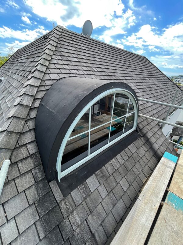 Architectural roofing: Close-up of a grey shingle roof with a curved window under a cloudy sky.